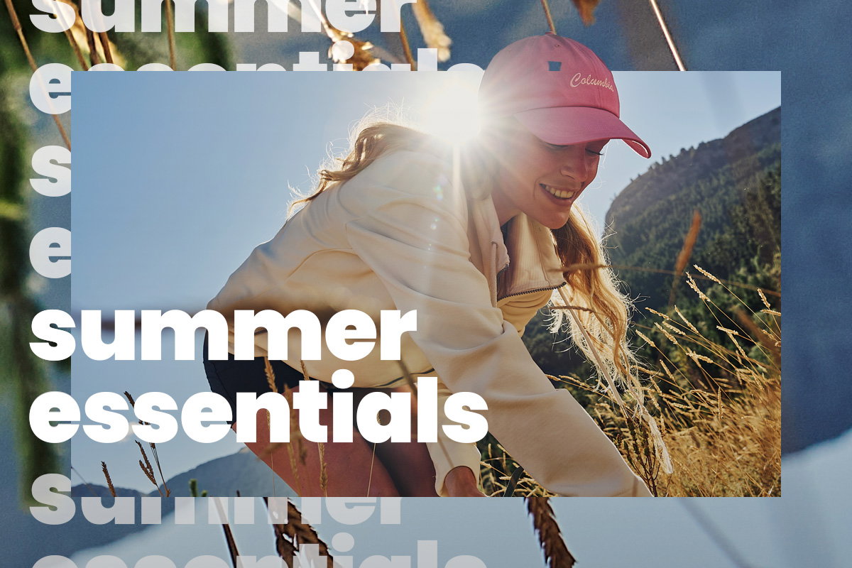 Person wearing a pink cap and light-colored jacket reaching through tall grass on a sunny hillside, with mountains in the background. Large overlay text reads “summer essentials.”