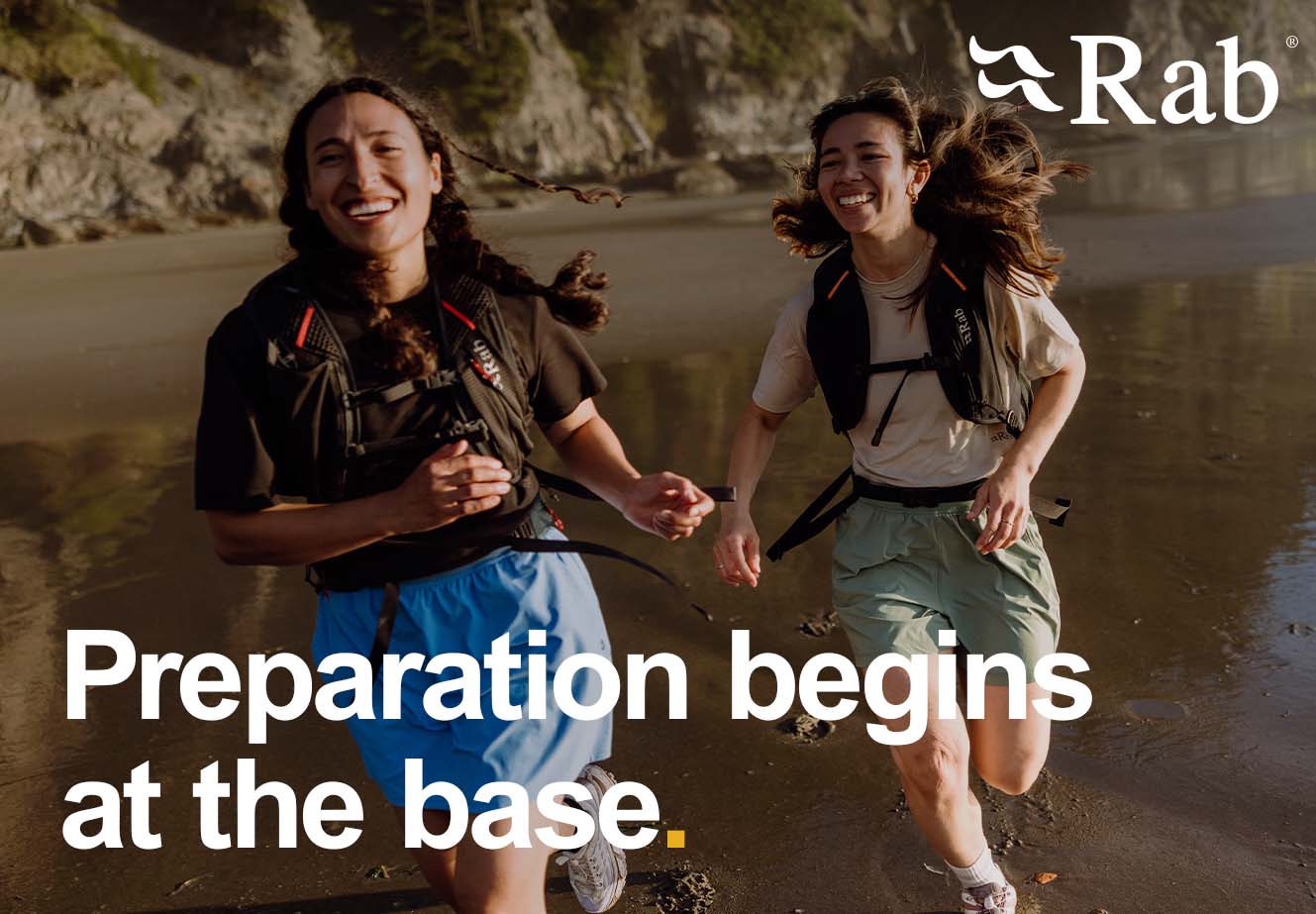 Two people wearing running gear and hydration packs jogging along a wet sandy shoreline with cliffs in the background. The Rab logo appears in the top right, and large overlay text reads “Preparation begins at the base.”