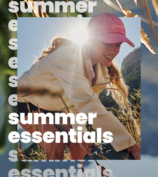 Person wearing a pink cap and light-colored jacket reaching through tall grass on a sunny hillside, with mountains in the background. Large overlay text reads “summer essentials.”
