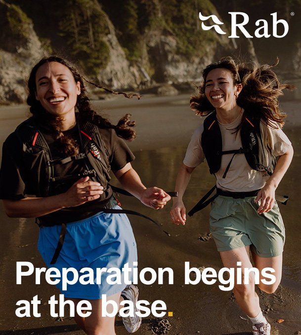 Two people wearing running gear and hydration packs jogging along a wet sandy shoreline with cliffs in the background. The Rab logo appears in the top right, and large overlay text reads “Preparation begins at the base.”
