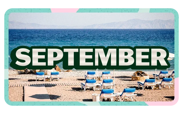 Turquoise border with pink corner accents, 'SEPTEMBER' white bold text with dark green outline, overcast grey-blue sky, distant mountain range, deep blue ocean, beige-tan pebbled beach, white sun loungers with blue cushions, dark beach umbrellas, beachgoers and wooden walkway.