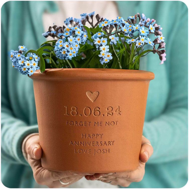 A terracotta plant pot engraved with a date, forget me not message and personalised dedication, held by someone wearing a teal jumper, planted with blue forget me not flowers