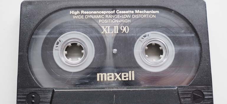 Close-up of a Maxell XL II 90 audio cassette tape with clear casing, showing the reels and tape inside. Labels include "High Resonanceproof Cassette Mechanism" and "Wide Dynamic Range + Low Distortion.