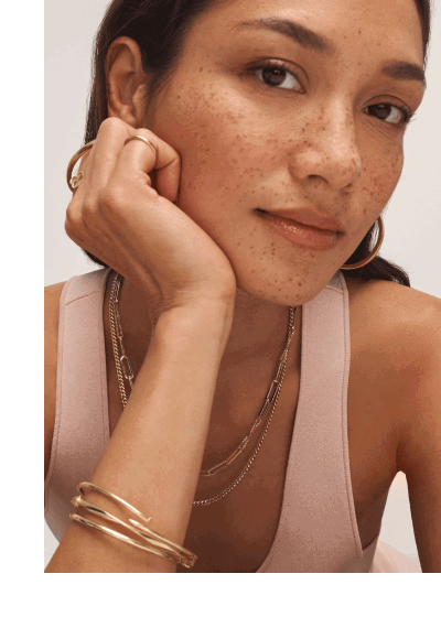 Close-up of a woman wearing layered yellow gold necklaces, hoop earrings, bracelets, and rings.