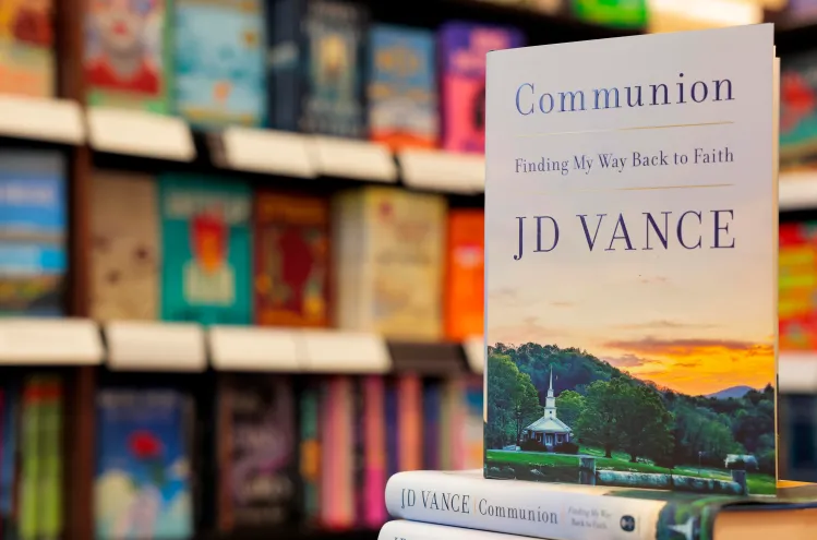 A copy of Communion by JD Vance standing on a bookstore table with shelves of other books in the background.