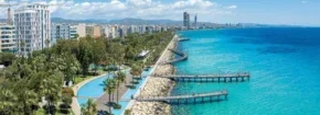 Palm-lined promenade along Limassol’s coastline with turquoise sea, a long pier stretching into the water, and city buildings lining the shore.