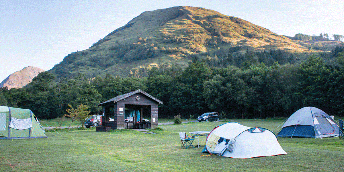 Camping site on a wide grassy field with several pitched tents of different sizes, including dome and family tents. A small wooden utility building sits near the centre, with parked cars nearby and camping gear such as folding chairs and a table set outside. Dense trees line the edge of the field, and a large sunlit mountain rises in the background under a clear sky.
