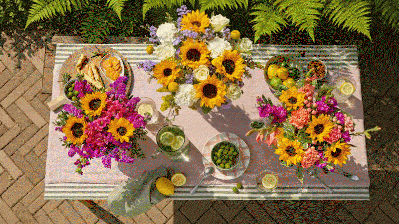 Vogelperspektive auf einen Gartentisch mit rosa Tischläufer, geschmückt mit drei sommerlichen Bouquets aus Sonnenblumen, weißen Rosen, Nelken, Levkojen und Meerlavendel. Dazu frische Limetten, Zitronen und Oliven für ein mediterranes Flair.