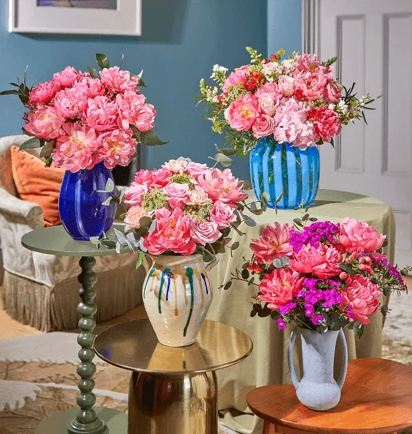Vier stilvolle Vasen mit üppigen, rosafarbenen Bouquets aus Coral Charm Pfingstrosen, Rosen und Löwenmäulchen. Sie stehen auf verschiedenen Beistelltischen vor einem hellblauen Hintergrund und verströmen sommerliche Eleganz.