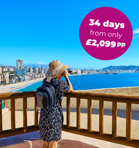 A person in a hat stands on a balcony overlooking a coastal city and blue sea.