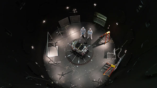 A birds-eye view of two people in white clean suits standing next to a spacecraft at the bottom of a large chamber. The pair are surrounded by ladders and other equipment. 