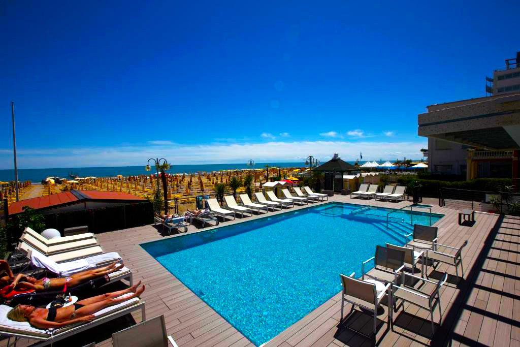 Rooftop hotel swimming pool with sun loungers and chairs overlooking a sandy beachfront and azure sea under a clear blue sky