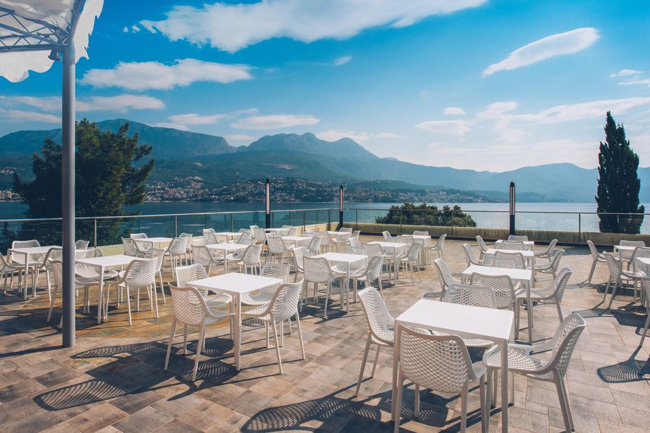 Empty outdoor restaurant terrace with white tables and chairs overlooking a calm lake and mountains under a blue sky