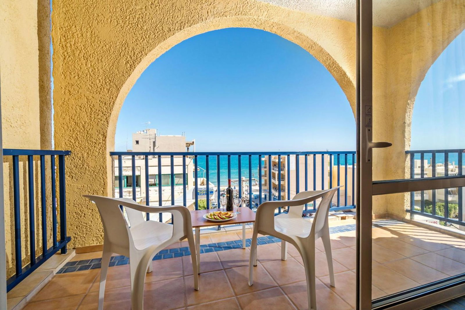 Balcony with two white chairs and small table under an arched yellow stucco opening, blue railing and sea view beyond nearby buildings
