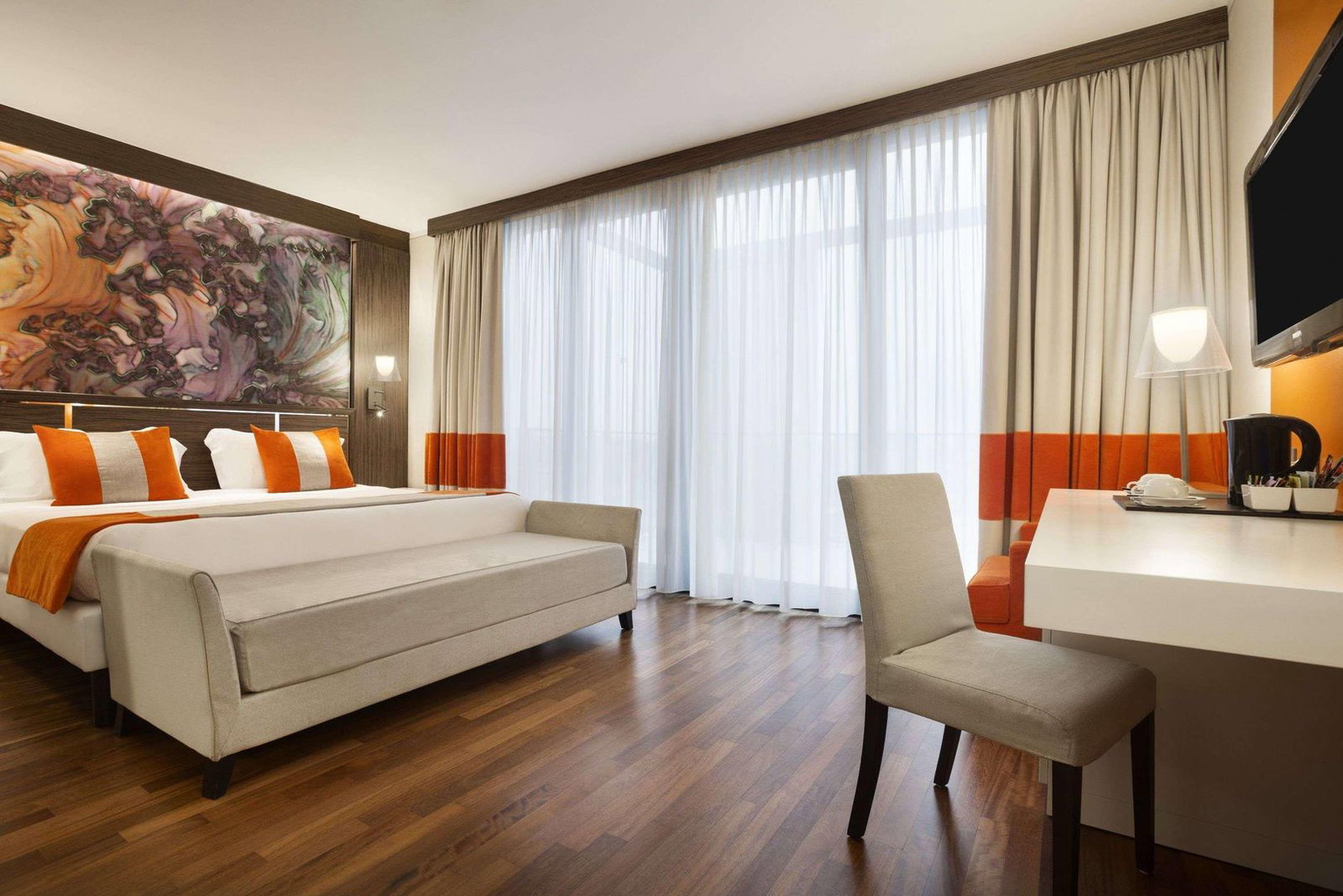 Modern hotel room with king bed, orange accent cushions and throw, upholstered bench, desk, wooden flooring and floor-to-ceiling curtains