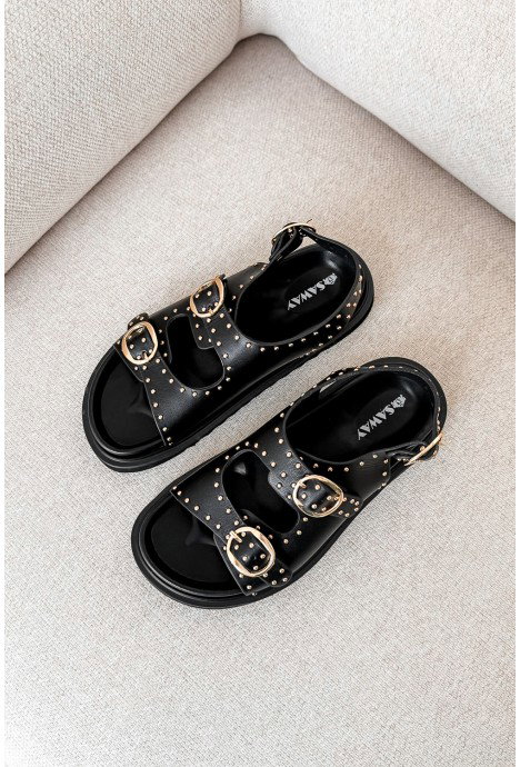 Pair of black studded leather sandals with three gold buckles on beige upholstered sofa