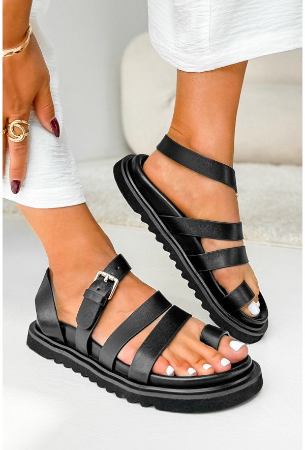 Black leather strappy platform sandals with buckle and chunky treaded sole on a model's feet, white pedicure and white dress