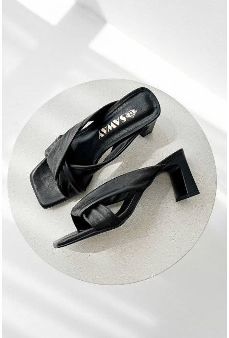 Top-down view of black leather crossover-strap block-heeled sandals on a round white table with soft sunlight shadows