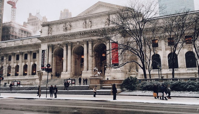 People walk in front of a large historic building with columns and statues during a snowy day.