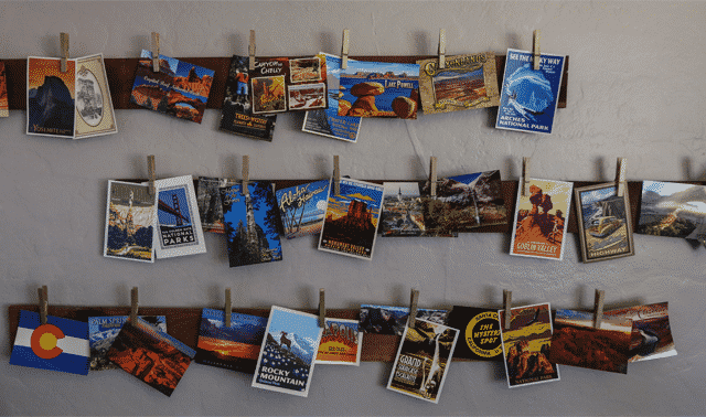 A display of various postcards and travel brochures clipped to two parallel strings across a gray wall. The postcards feature images and names of different national parks and scenic destinations.