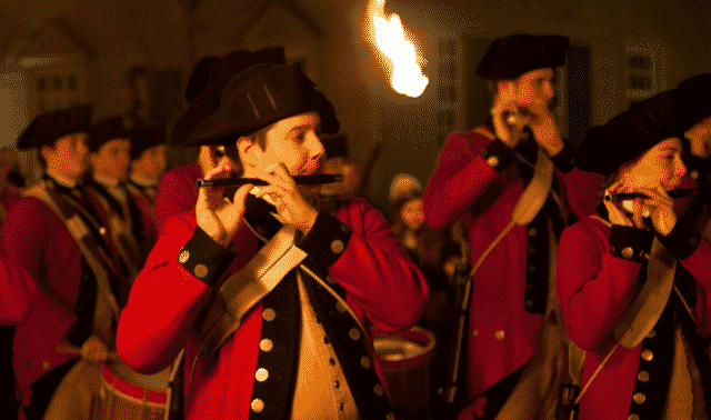 A group of people dressed in historical military uniforms are playing flutes. One member is holding a torch.