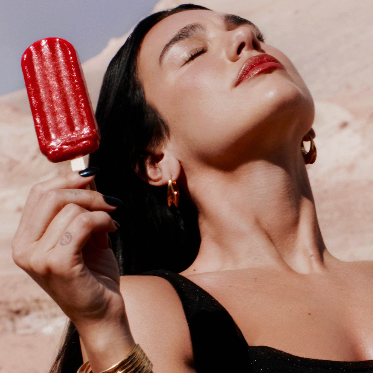 Singer Dua Lipa tilts her head back in the sun, wearing gold earrings and holding a red popsicle.