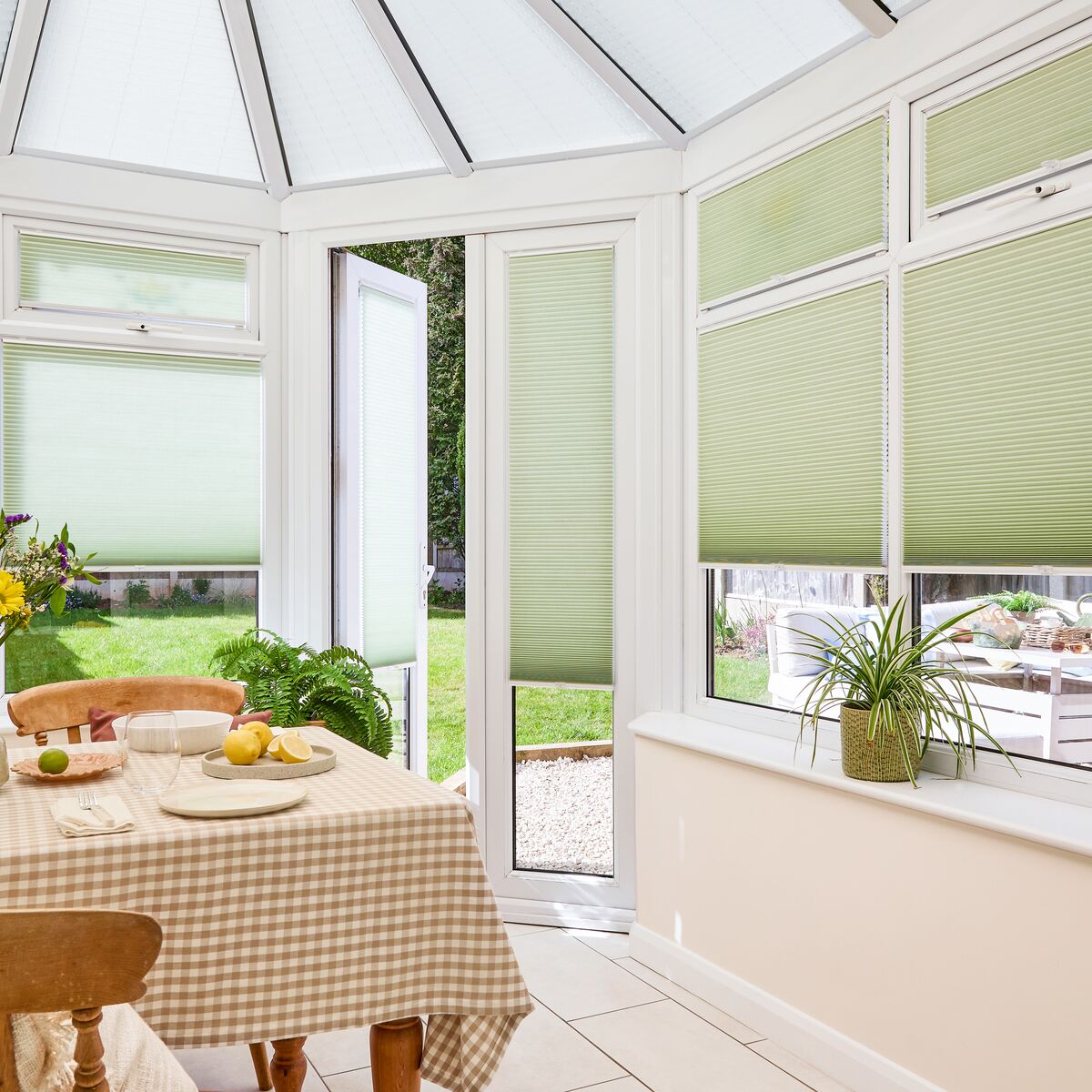 Multiple sage green honeycomb blinds installed across the glass panels of a bright conservatory dining area