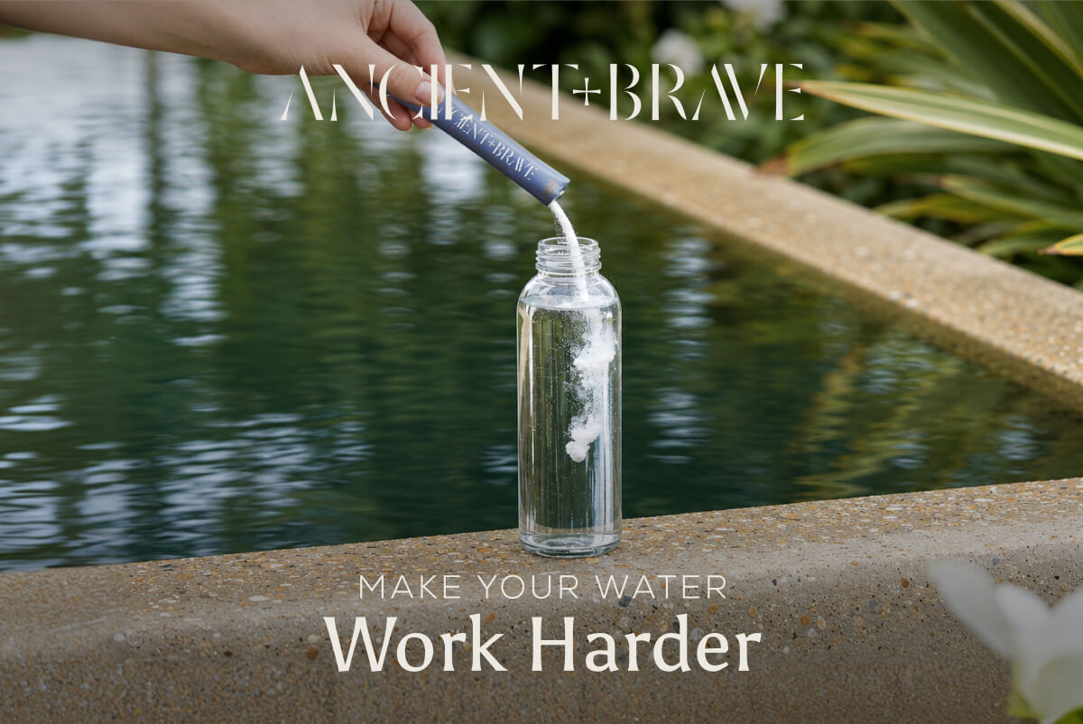 Ancient + Brave. A hand pours a sachet of powder into a glass water bottle. Make Your Water Work Harder.