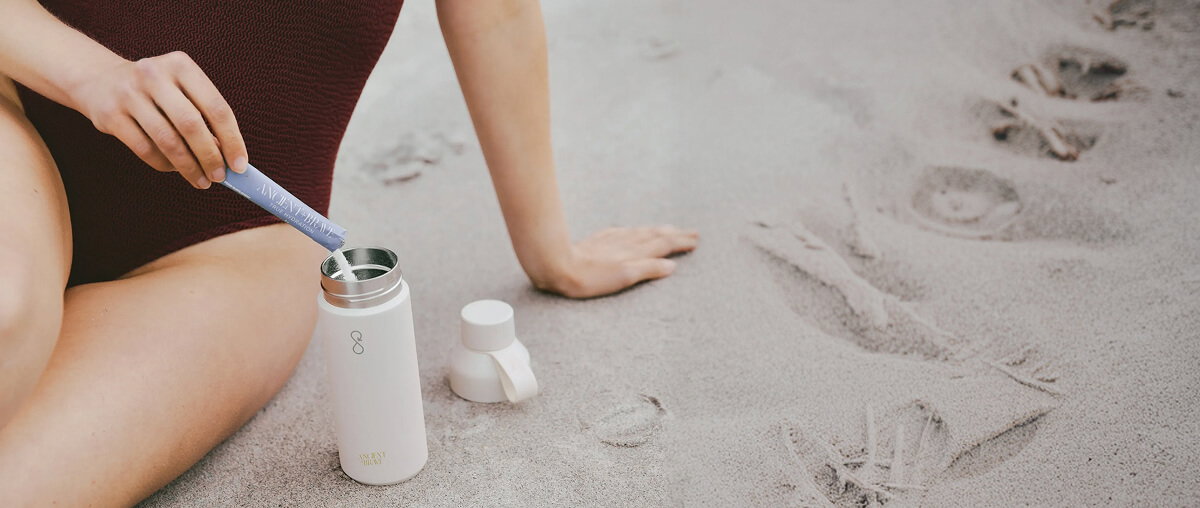 A close up of a person sitting on sand pouring a sachet of an Ancient and Brave supplement into a water bottle