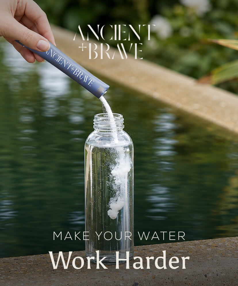 Ancient + Brave. A hand pours a sachet of powder into a glass water bottle. Make Your Water Work Harder.