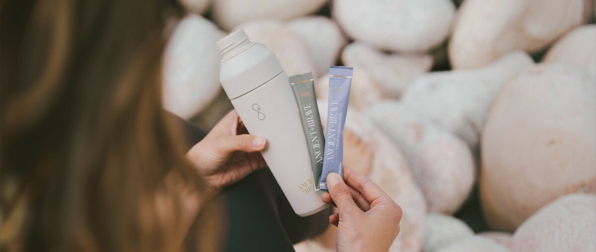 A woman holding a water bottle and two sachets of Ancient + Brave