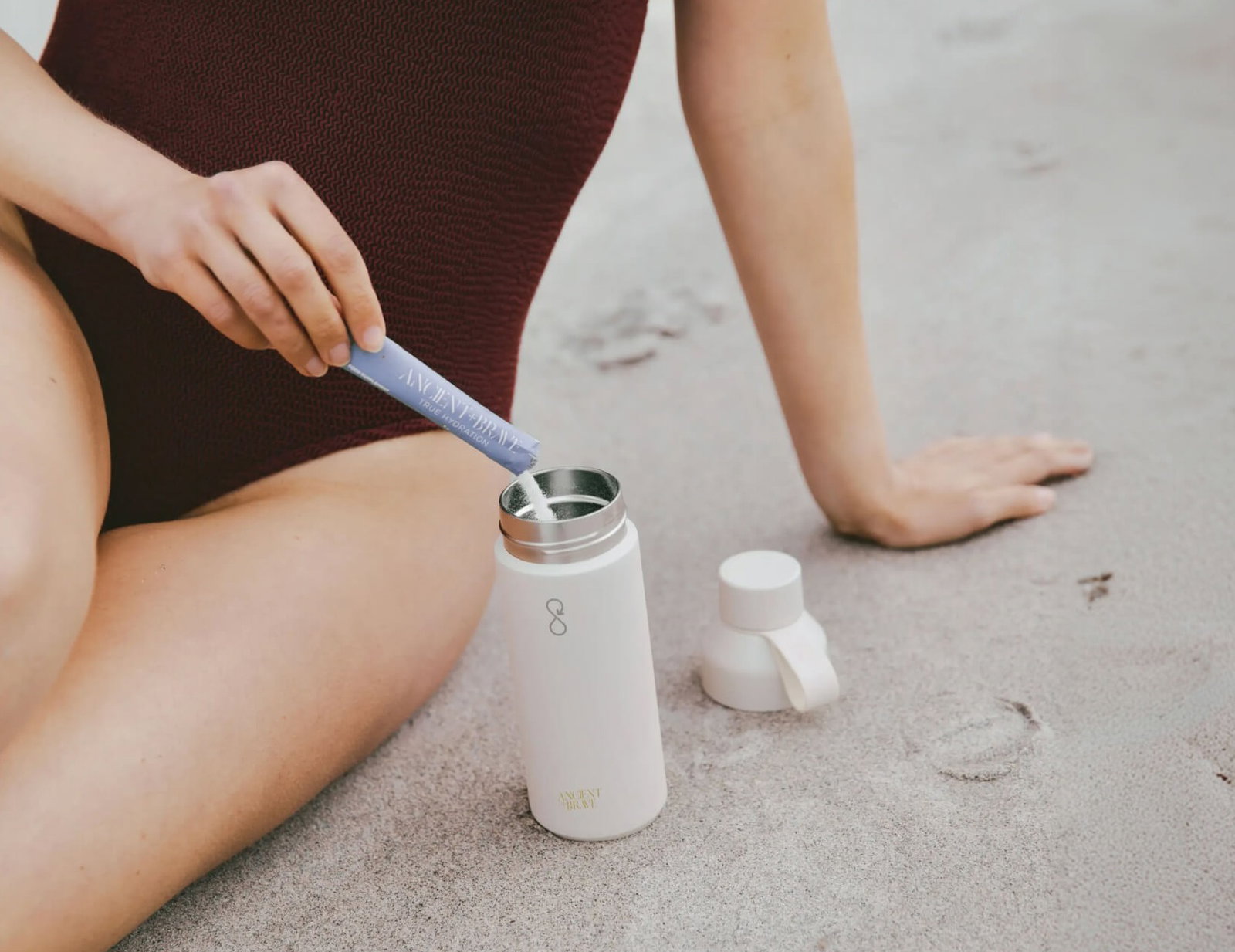A close up of a person sitting on sand pouring a sachet of an Ancient and Brave supplement into a water bottle