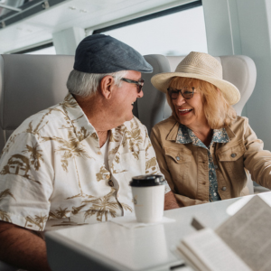 An older couple sits together on a modern train, engaged in conversation. They wear casual vacation clothing, including hats and light jackets, suggesting a relaxed trip.