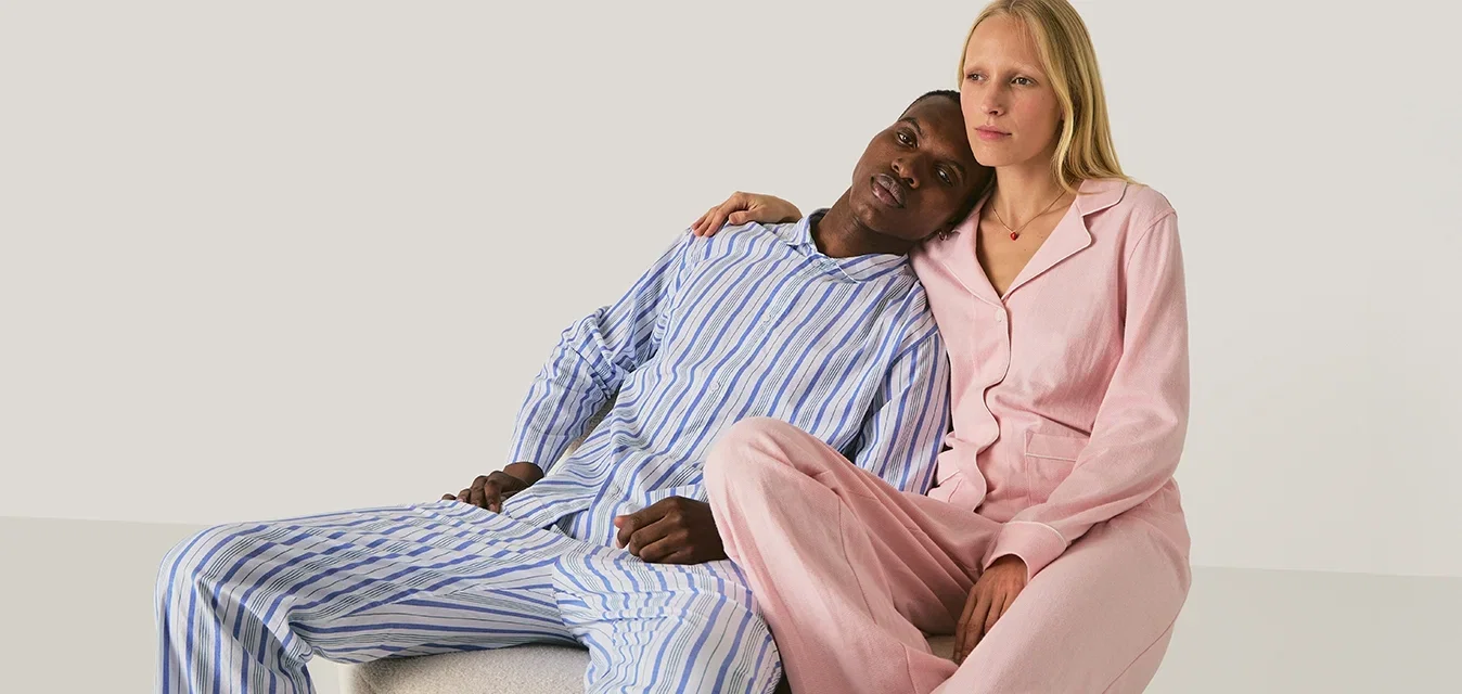 Ein Mann in blau gestreiften Pyjamas legt seinen Kopf auf die Schulter einer Frau, die rosa Pyjamas trägt, und sitzt dicht nebeneinander auf einem beigen Block.