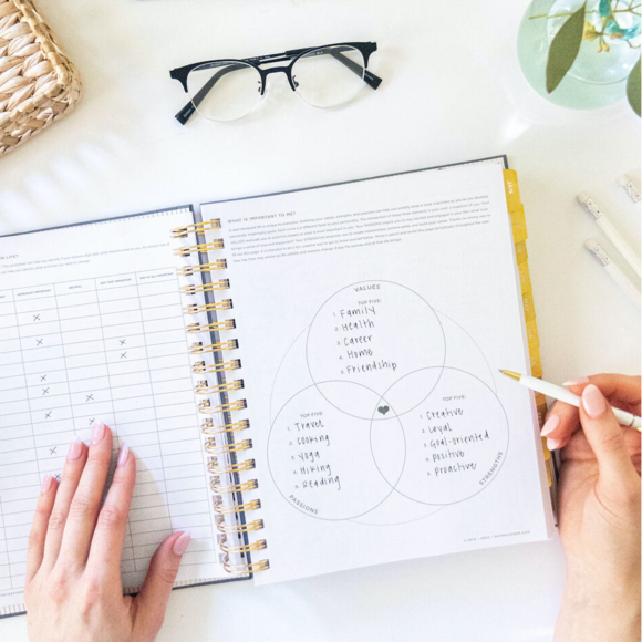 hands holding open book planner showing self-assessment worksheets, glasses, vase, pens