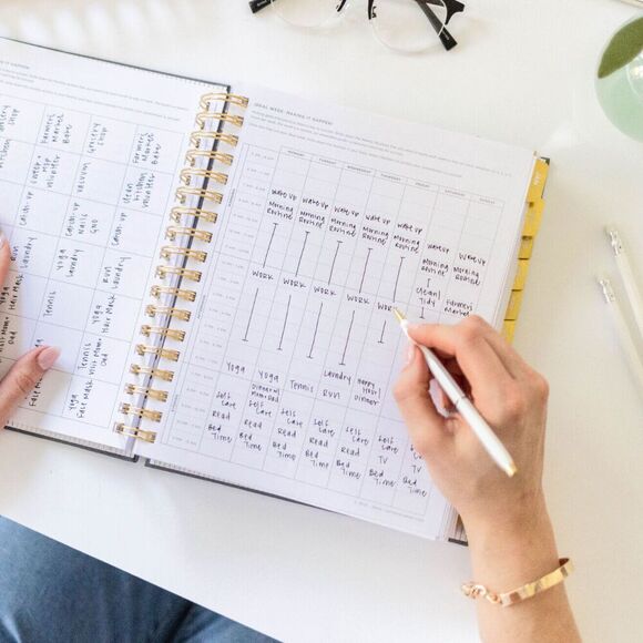 hands holding open book planner showing ideal month and week worksheets, glasses, vase pens