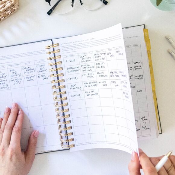 hands holding open book planner showing goal-setting worksheets, glasses, pen