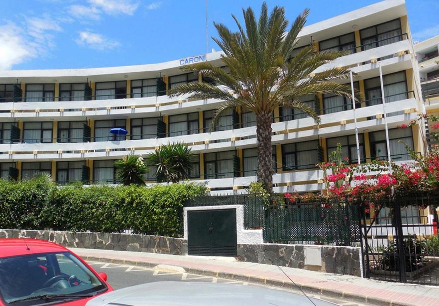 Curved white hotel with rows of balconies, central palm tree and bougainvillea-lined fence, parked cars on sunlit street