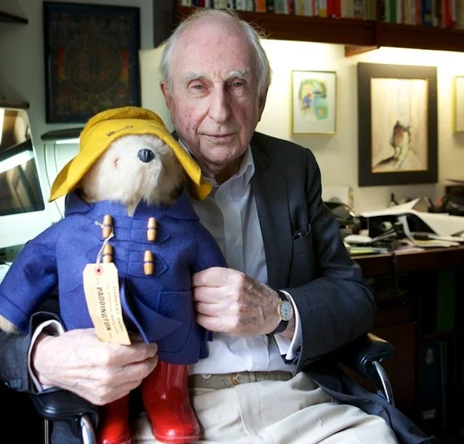 The late Michael Bond, the author who created Paddington Bear. He is seated in his study holding a large Paddington soft toy. Paddington is wearing his trademark floppy hat, blue duffle coat and red Wellington boots.