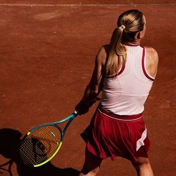 Female tennis player with blonde ponytail hitting a backhand on red clay court, wearing white top and red skirt and holding a racket