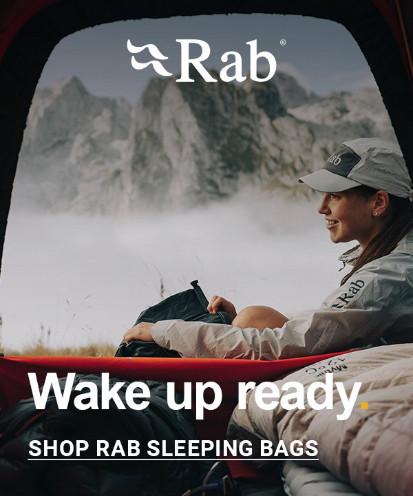 Outdoor scene viewed from inside a tent showing a person sitting beside a sleeping bag and pillow, looking out over a misty mountainous landscape. The person is wearing a light-colored jacket and cap branded “Rab.” The background features rugged rocky peaks rising above a layer of low cloud.