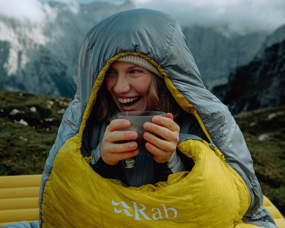 Close-up outdoor scene of a person wrapped in a hooded sleeping bag, holding a cup with both hands while sitting in a mountainous landscape. The sleeping bag is grey with a yellow lining and displays the “Rab” logo on the front. In the background, rugged mountains and cloudy skies create a cool, alpine setting, with muted greens and greys suggesting high-altitude terrain.