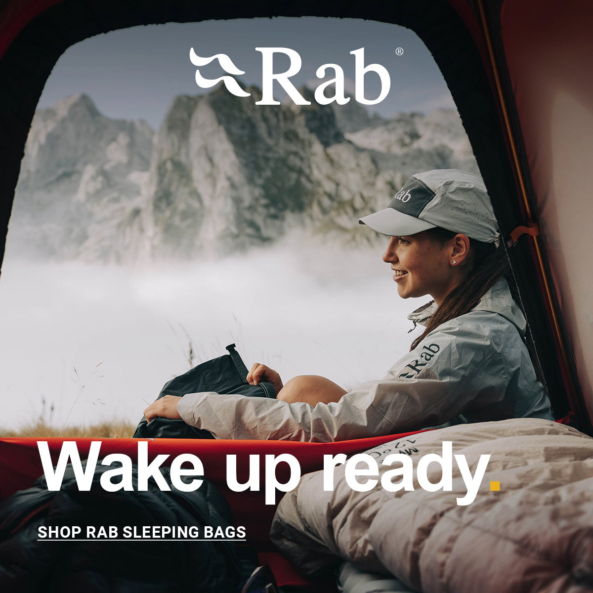 Outdoor scene viewed from inside a tent showing a person sitting beside a sleeping bag and pillow, looking out over a misty mountainous landscape. The person is wearing a light-colored jacket and cap branded “Rab.” The background features rugged rocky peaks rising above a layer of low cloud.