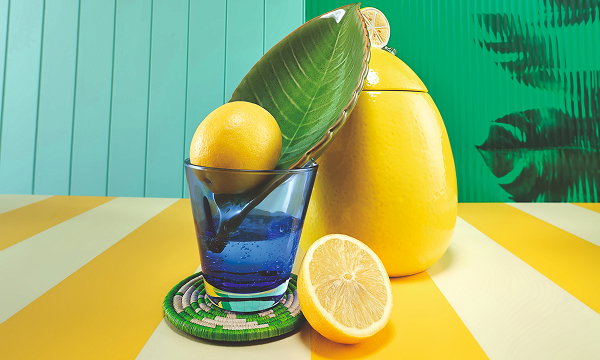 On a yellow striped surface sits an egg-shaped yellow pot, a blue glass, lemons, a green leaf dish, and a coaster. The background is a green wall.