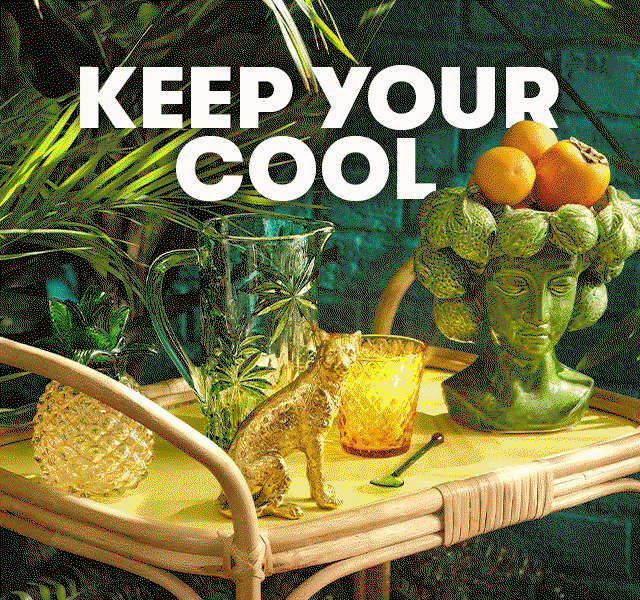 Tropical table scene with green glassware, a gold animal figurine, a leafy fruit bowl with oranges, and lush plants against a teal brick background.