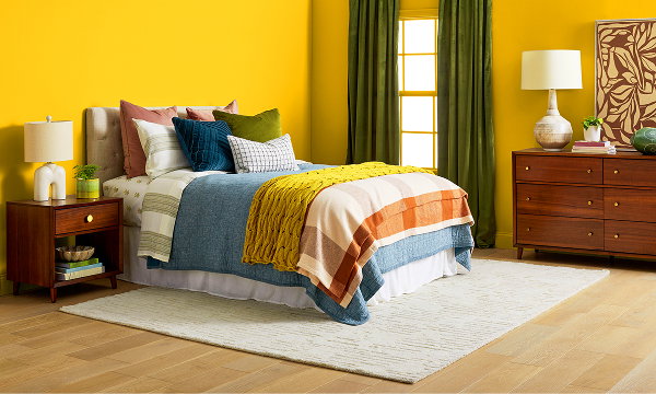 Bedroom with a yellow wall, green curtains, wooden side tables and matching chest of drawers, light rug, and bed styled with colourful sheets.