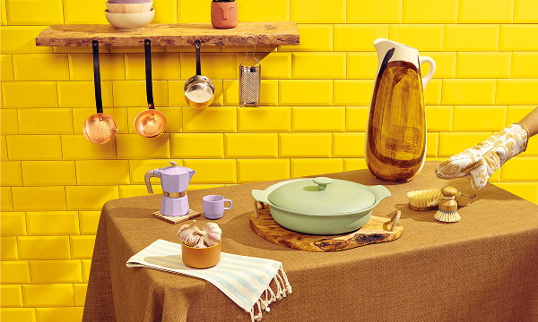 Kitchen scene with colourful cookware, a pastel coffee maker, hanging copper pans, a tall pitcher, and a pot on a brown table against a yellow wall.