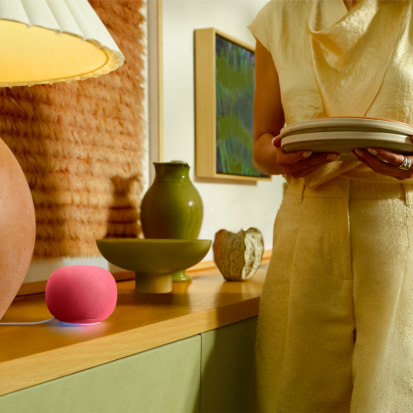 Pink HomePod mini on a wooden sideboard beside green vases and a person holding plates.