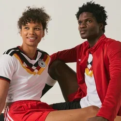Zwei junge Fussballspieler in Teamuniformen sitzen auf dem Boden, einer hält einen Fussball, vor einem schlichten weissen Hintergrund.