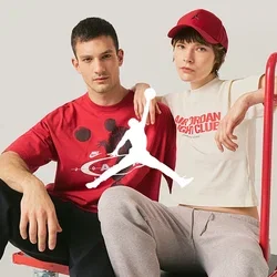 Zwei junge Erwachsene sitzen auf einem roten Flachwagen; einer trägt ein rotes T-Shirt mit Grafik, der andere ein weißes bauchfreies T-Shirt und eine rote Kappe.